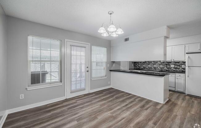 A modern kitchen and dining area featuring a pendant light, large windows with natural light, and a sliding glass door. The kitchen includes sleek cabinets and a backsplash with a mosaic design. The flooring is wood-like, adding warmth to the space.