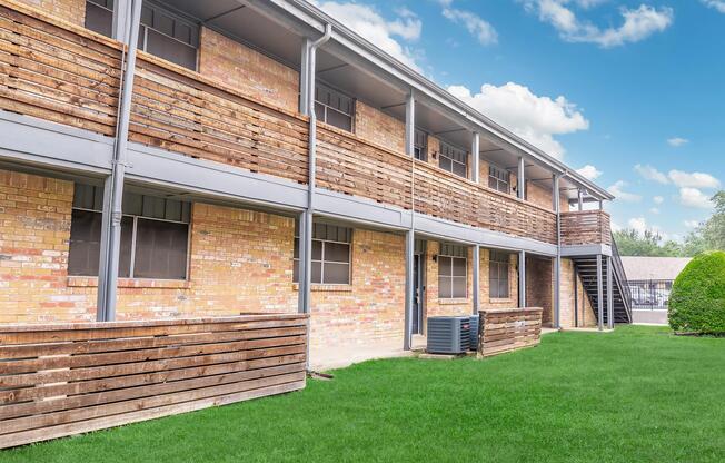 A two-story brick building with wooden balcony railings and grassy landscaping. The structure features multiple windows and a staircase on one side, against a backdrop of a blue sky with scattered clouds.