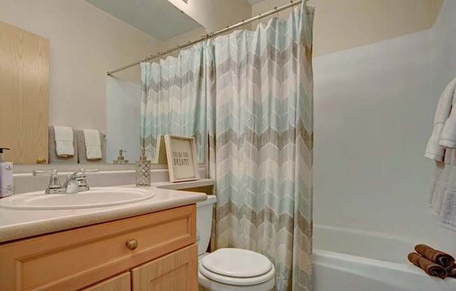 A welcoming bathroom here at Aspen Creek featuring a light wood vanity with a white countertop and sink, a wide wall mirror with neatly folded towels, and a shower and tub combination framed by a patterned fabric curtain, complemented by neutral wall tones, simple décor accents, and soft lighting that creates a clean and comfortable atmosphere.