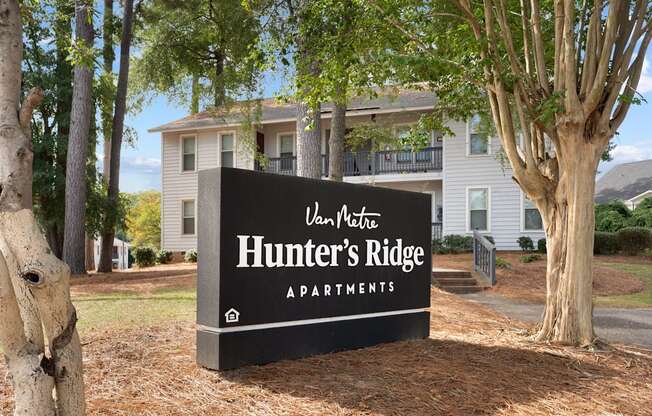 A sign for Hunter's Ridge Apartments is in front of a tree and a house.