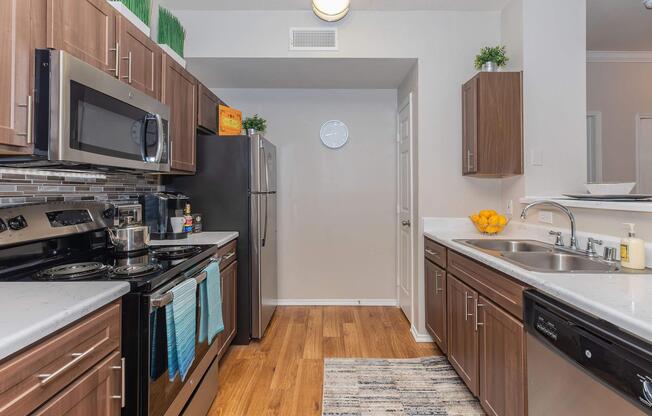 A modern kitchen featuring stainless steel appliances, including an oven, microwave, and refrigerator. The countertops are light-colored with a dark wood cabinet finish. A rug is placed on the wooden floor, and there are decorative plants and oranges on the counter for a cozy touch.