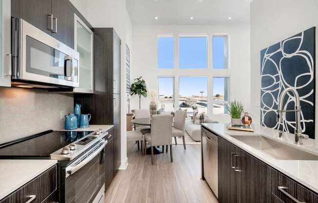 1 Bedroom Apartments in Woodland Hills, CA - The Q Variel - Modern Kitchen with Custom Cabinets, Quartz Countertops, Luxury Wood Plank Style Flooring, Stainless Steel Appliances, Large Kitchen Island, High Ceilings, and Open-Concept Layout with Dining Area and Living Room Boasting Expansive Windows