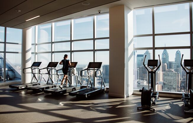 A person is exercising on a treadmill in a brightly lit gym with a view of the city.