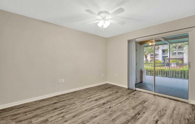 A spacious, well-lit room featuring a ceiling fan, with light-colored walls and hardwood-style flooring. A sliding glass door leads to a porch area with outdoor views, creating a welcoming and airy atmosphere.