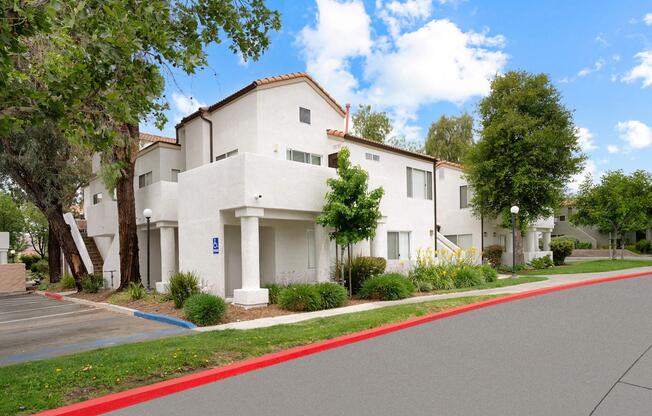 A modern residential building with multiple white stucco apartments surrounded by lush greenery. The scene features a clear blue sky with a few clouds, decorative yellow flowers, and a red-bordered path leading to the entrance. Parking spaces are visible nearby.