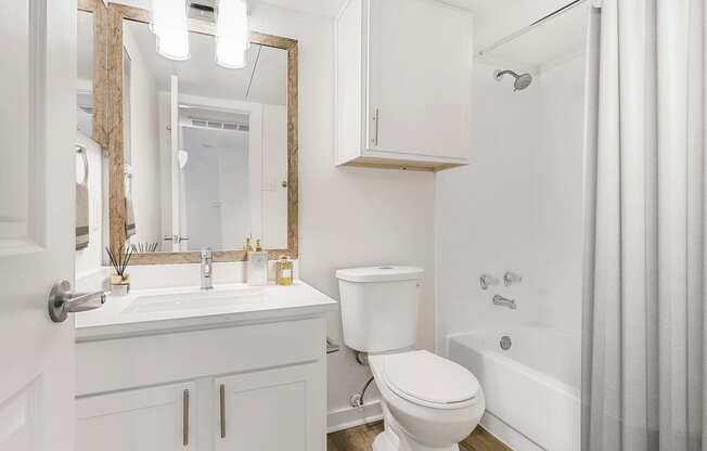 Virtually staged bathroom with white cabinetry, white quartz countertop, custom vanity mirror, medicine cabinet, wood style flooring, sheer shower curtain and toilet