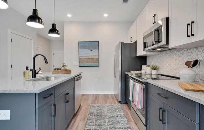 Modern kitchen featuring grey and white cabinetry, stainless steel appliances, and a geometric backsplash. A large island with a sink is positioned in the center. Pendant lights hang above, and a decorative rug is laid on the floor. Wall art adds a touch of color. The space is well-lit and organized.