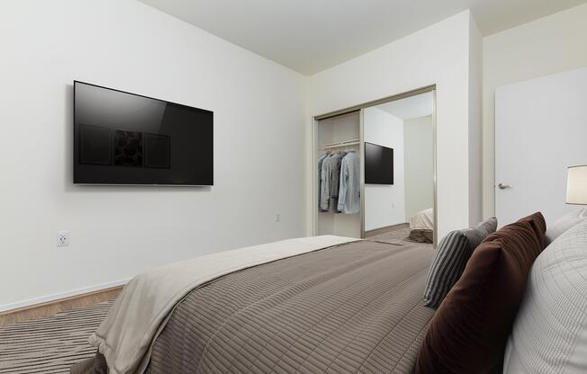 A modern bedroom featuring a neatly made bed with a light brown quilt and decorative pillows. There is a large flat-screen TV mounted on the wall opposite the bed. A mirrored closet door reflects the room, which also shows a few items of clothing. The walls are painted a soft color, creating a calm atmosphere.