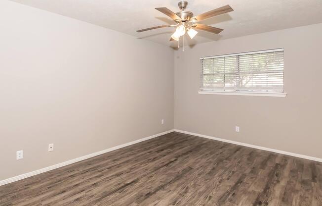A spacious, empty room with light gray walls and a ceiling fan. The floor features wood-like laminate, and there is a window with blinds allowing natural light. The overall atmosphere is bright and airy, ideal for decorating or furnishing.