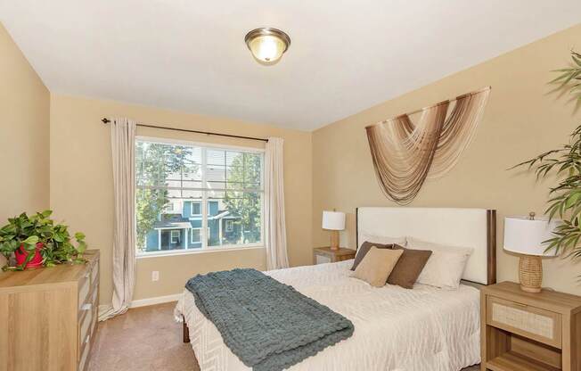 A bedroom with a large bed and a view of a blue townhome through the window at The Landing Apartments and Townhomes, Oregon, 97045
