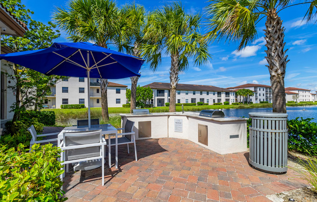 A patio with a table and chairs under an umbrella with a trash can and palm trees.