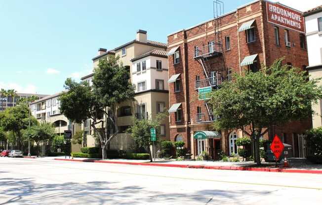 Street view of Brookmore apts in Pasadena CA. Four story brick building with "now leasing" sign and large green trees and bushes in front