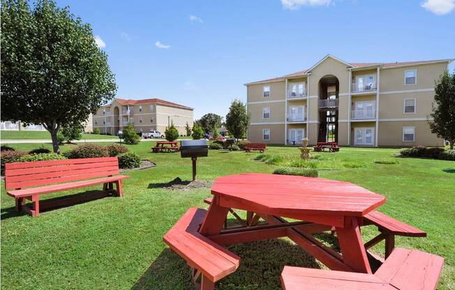 A red picnic table is in the foreground of a grassy area with benches and a building in the background.