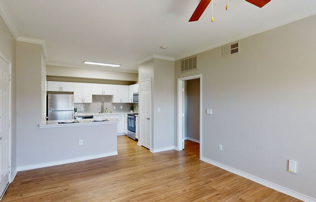 A living and kitchen area here at Landing at Mansfield with an empty open layout, warm wood floors, white kitchen cabinets, breakfast bar, and soft neutral walls with bright overhead lighting.