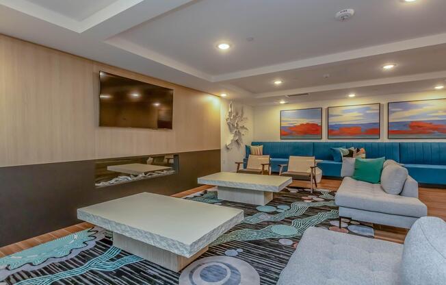 A modern lounge area featuring a light gray sofa and two matching coffee tables on a decorative rug. The walls are adorned with abstract artwork in warm colors. A flat-screen TV is mounted on one wall, while a blue upholstered bench provides additional seating along another wall. Soft lighting creates a cozy atmosphere.