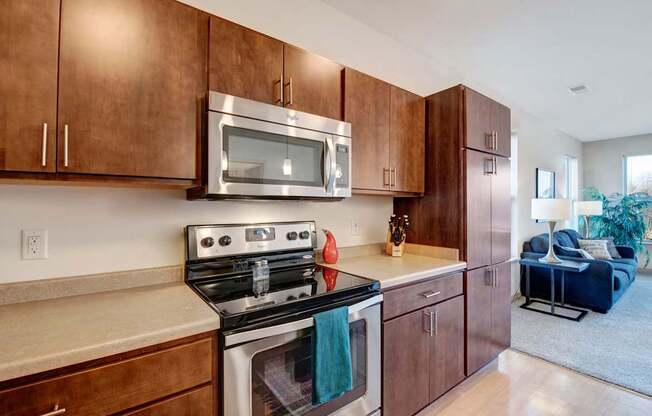 A modern kitchen here at Vue at Pinnacle Park with rich wood cabinets, stainless steel stove and microwave, smooth countertops, and generous storage, opening toward a bright, comfortable living area with soft carpet and natural light.