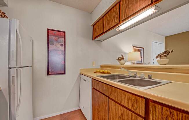 A compact kitchen here at Cypress Pointe with warm wood cabinets, beige countertops, and a double stainless steel sink beneath bright overhead lighting. White appliances, including a refrigerator and dishwasher, add a clean, functional feel to this cozy, practical cooking space.