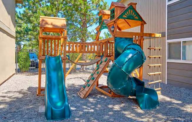 A playground with two slides, a swing set, and a small rock climbing wall surrounded by pea gravel and trees in the background.at Wildflower, Washington
