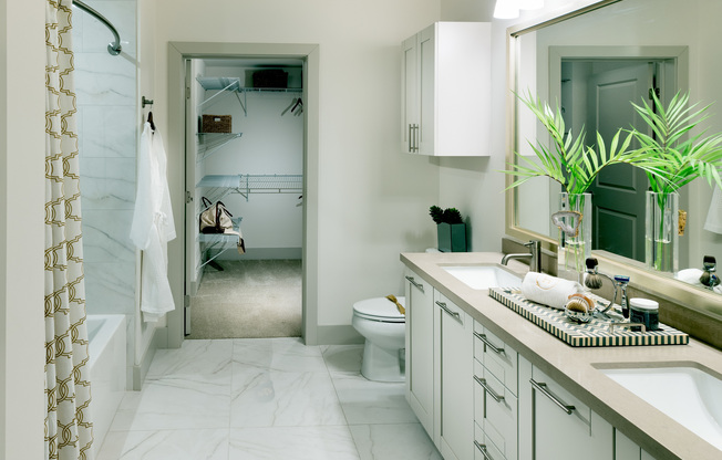 A bright bathroom with a dual vanity, large mirror, white cabinets, a large tub, marble tile on the floor and bathtub walls, and a walk-in closet.