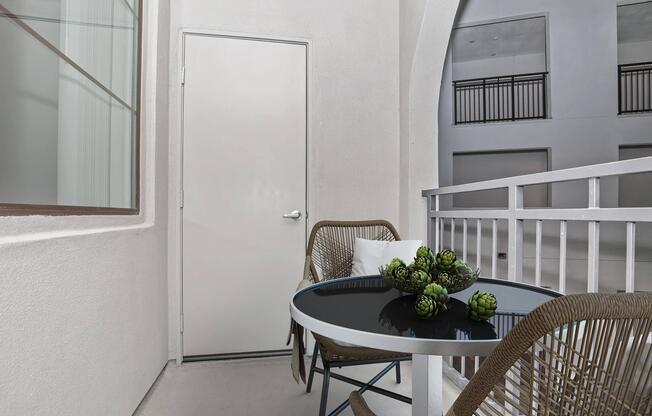 A minimalist balcony setup featuring a round table with a black surface, surrounded by two wicker chairs. A potted arrangement of green succulents sits on the table. The background shows a plain white wall and a closed door, with a subtle view of an interior space.
