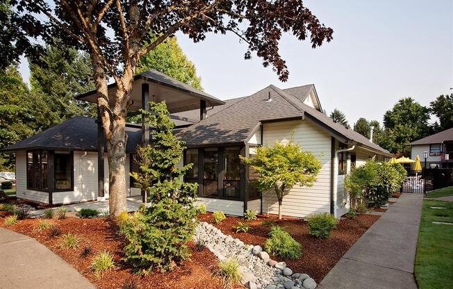 A house with a white siding and a brown roof is surrounded by a garden with a variety of plants and trees.