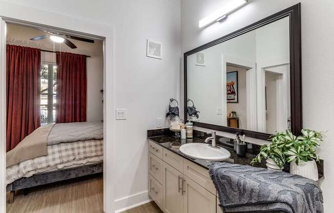 A bathroom with a bedroom door open to a bedroom with red curtains.