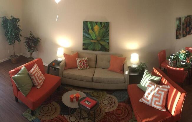 A cozy living room featuring a light grey sofa with colorful cushions, two bright orange chairs with patterned pillows, a circular coffee table, and a decoratively patterned rug. The space includes several indoor plants, warm lighting from lamps, and colorful artwork on the walls.