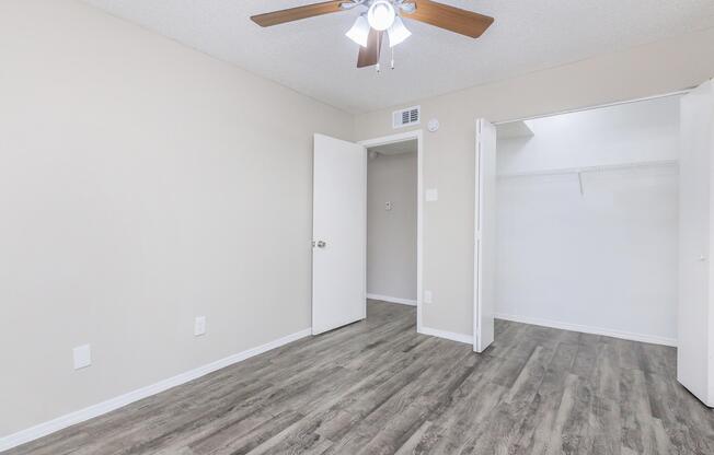 A light-colored room with a ceiling fan, featuring beige walls and a wooden floor. There are two white doors, one leading to an empty closet with a rod for hangers. The room has ample natural light, creating a bright and airy atmosphere.
