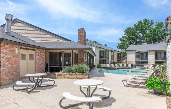 Outdoor pool area with lounge chairs, a stone wall, and picnic tables. Surrounding buildings feature a mix of brick and siding, with greenery adding to the ambiance. The sky is bright blue, indicating a sunny day.
