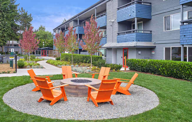 Exterior view of the building and community area. Outdoor area w/ brick fireplace with 6 orange lawn chairs around it. Fire pit space is surrounded by green grass with a path leading up to it.