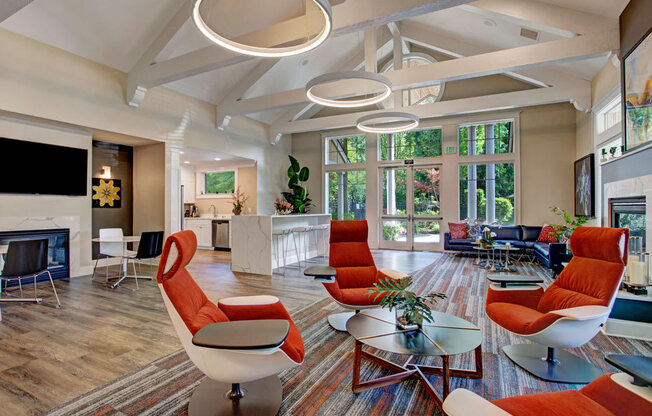 A spacious resident lounge here at Portsmith featuring soaring vaulted ceilings with exposed beams, modern circular pendant lighting, and large windows that frame lush outdoor views, with contemporary seating in warm accent tones, a fireplace with mounted television, wood-style flooring, and an open kitchen area with a large island that creates an inviting space for relaxing or gathering.