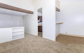 Bright and spacious interior of an apartment featuring beige carpet flooring, light-colored walls, and a small kitchen area visible in the background. There is a partitioned space with built-in shelves, and a bar counter connecting to the kitchen. Natural light enhances the open layout and modern feel of the space.
