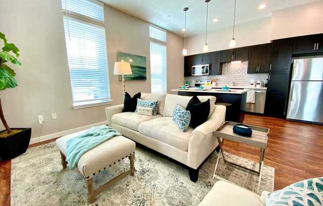 A living room with hardwood style flooring with two windows with open access to a kitchen with an island bar and hanging pendant lights.