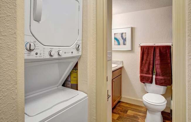 A convenient in-home laundry and bathroom area here at Heritage Hills featuring a stacked washer and dryer tucked into a hallway alcove, adjacent to a bathroom with wood-style flooring, a vanity with countertop sink, framed wall art, and a toilet accented by neatly hung towels for a clean, functional layout.