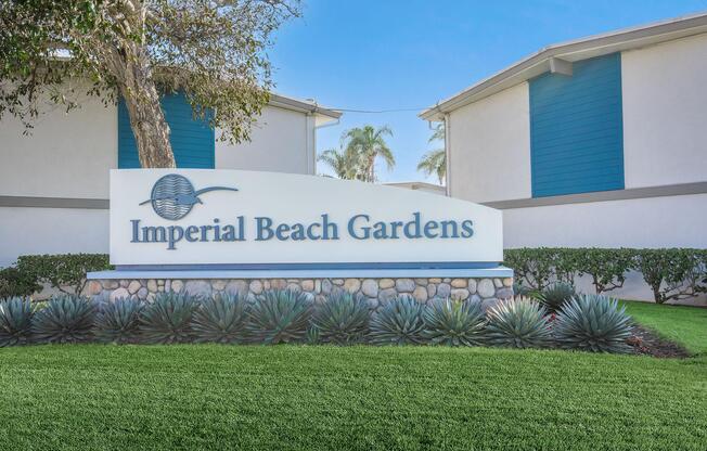 Sign for Imperial Beach Gardens, featuring the name prominently displayed. The sign is surrounded by landscaped greenery, including low shrubs and decorative stonework, with a clear blue sky in the background.