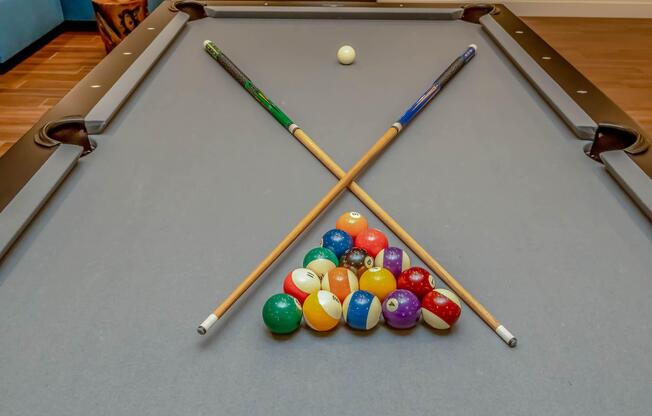 A pool table with a triangle rack of colorful billiard balls arranged at the bottom and two pool cues crossed above the rack. A single white cue ball rests on the table, ready for the game to begin. The background features a softly lit room.