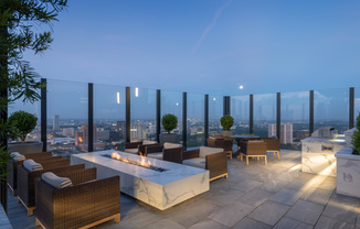 Multiple seating areas placed at the corner of the rooftop with glass walls allowing for a panoramic view of the city. There are 6 armchairs around a minimalist marble fire pit, a patio table and chairs, and landscaping throughout.