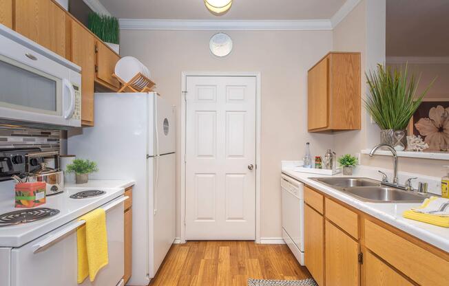 A clean and modern kitchen featuring wooden cabinets, white appliances including a microwave, refrigerator, and dishwasher. The countertop is equipped with a sink, and there are small plants and decorative items. A clock is mounted on the wall, and a door leads to another room. The floor is made of warm-colored wood.
