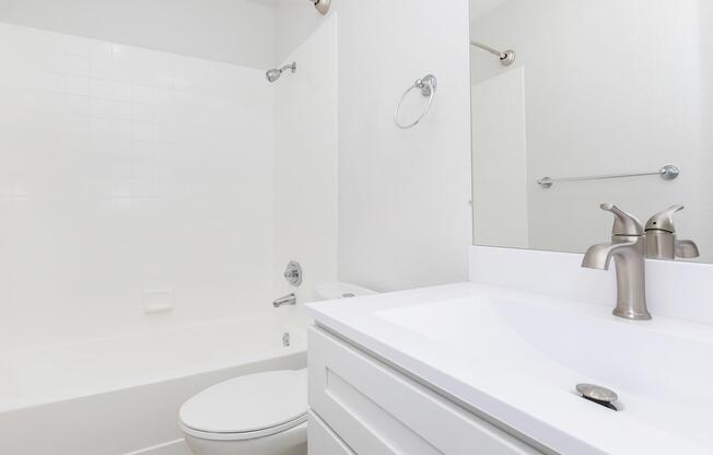 A clean and modern bathroom featuring a bathtub, shower combination, a white countertop with a faucet, and a toilet. The walls are painted white, and there is a mirror reflecting the space. A towel bar is mounted on the wall, adding to the bathroom's functionality and sleek design.