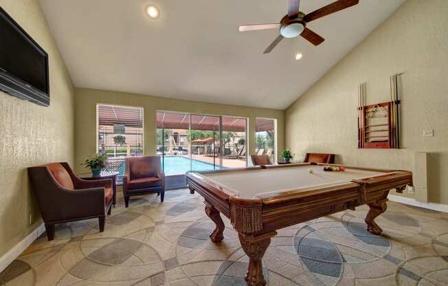 A bright game room here at Trinity Place featuring a full-size pool table centered on patterned carpet. Comfortable lounge chairs line the walls, and a mounted television adds entertainment. Large windows and glass doors overlook the pool area, while high ceilings with ceiling fans and recessed lighting create an open, inviting community space.