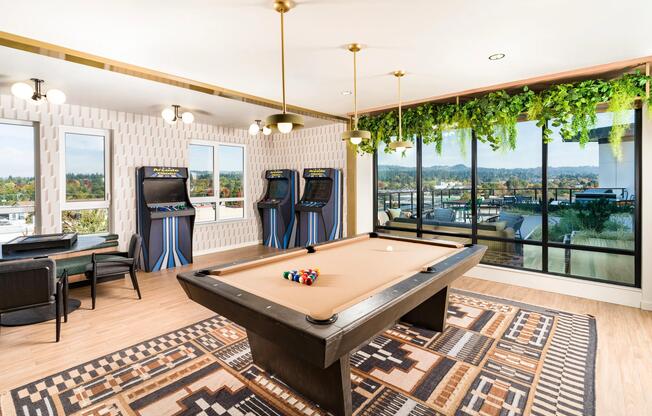 A game lounge at Modera Beaverton with a pool table, arcade games, and large windows that bring in daylight and views beyond the room.