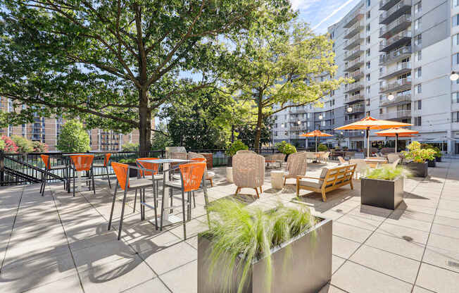 A patio with orange chairs and tables is surrounded by trees and buildings.