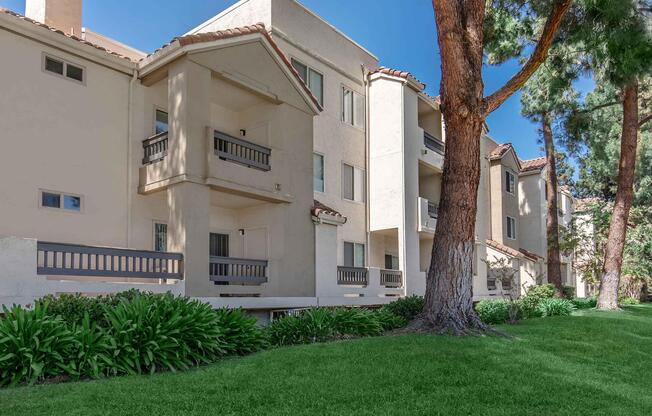 A view of a residential apartment community featuring multiple white stucco buildings with balconies. Lush green grass and shrubs line the foreground, and tall trees provide shade nearby. The architecture reflects a modern design with Spanish influences, set against a clear blue sky.
