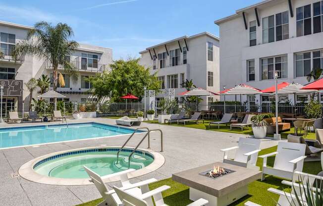 A pool with a hot tub and chairs in the foreground with apartment buildings in the background.