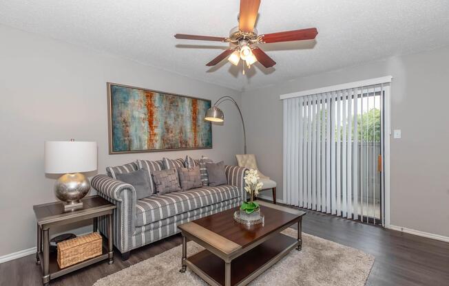 A modern living room featuring a striped sofa with decorative pillows, a stylish floor lamp, and a coffee table with a plant centerpiece. There's a large piece of abstract wall art above the sofa, and vertical blinds cover a sliding glass door leading to an outdoor space. The flooring is dark wood, and a rug adds warmth to the area.