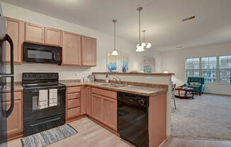A bright kitchen here at Swan Creek with warm wood cabinets, black appliances, and open breakfast bar. Inviting layout flows into a cozy living room with teal accent chair.