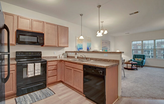 A bright kitchen here at Swan Creek with warm wood cabinets, black appliances, and open breakfast bar. Inviting layout flows into a cozy living room with teal accent chair.