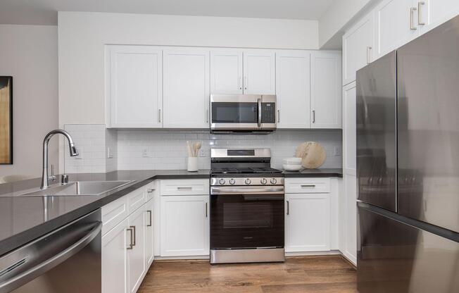 Modern kitchen featuring white cabinets, a stainless steel gas range with a microwave above, a silver refrigerator, and a dark countertop. The space includes a sink, wooden cutting board, and decorative kitchenware.