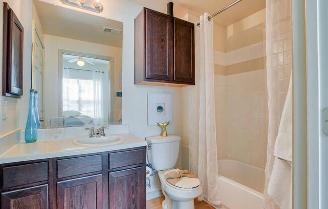 A bathroom with a toilet, sink, side wall mirror with storage, wood cabinets, wood floor and bathtub