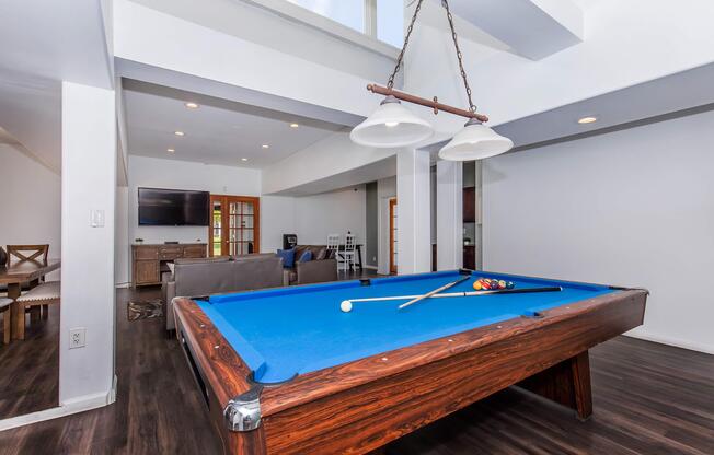Interior shot of a spacious game room featuring a blue felt pool table with a cue and balls. The room is well-lit with recessed lighting and a large window, and there are modern furnishings including a sofa and a television in the background. The flooring is dark wood, creating a warm atmosphere.